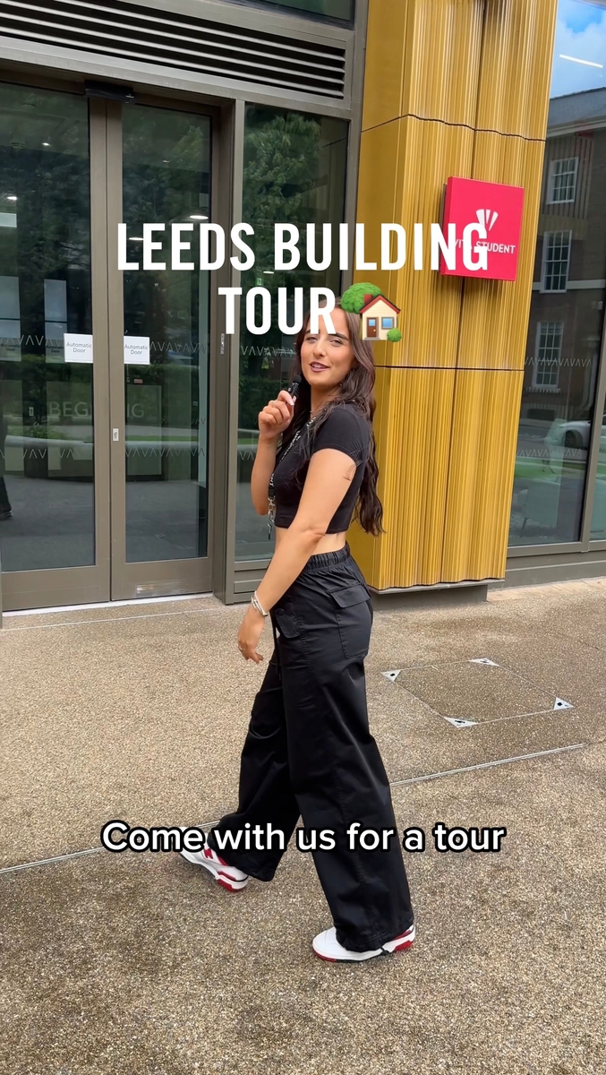 A person in black clothing stands outside a building with glass doors, looking back and smiling. The text reads "Leeds Building Tour" with a house emoji and "Come with us for a tour." The surroundings include reflective windows and a pink sign.