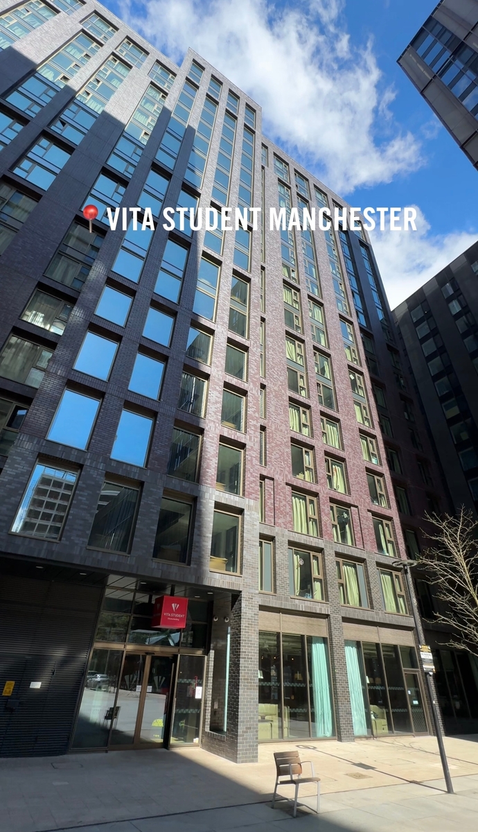 Tall modern building with numerous windows and a small entrance canopy. A sign reads "VITA STUDENT MANCHESTER" at the top. The street is empty with a chair in the foreground and a clear blue sky above.