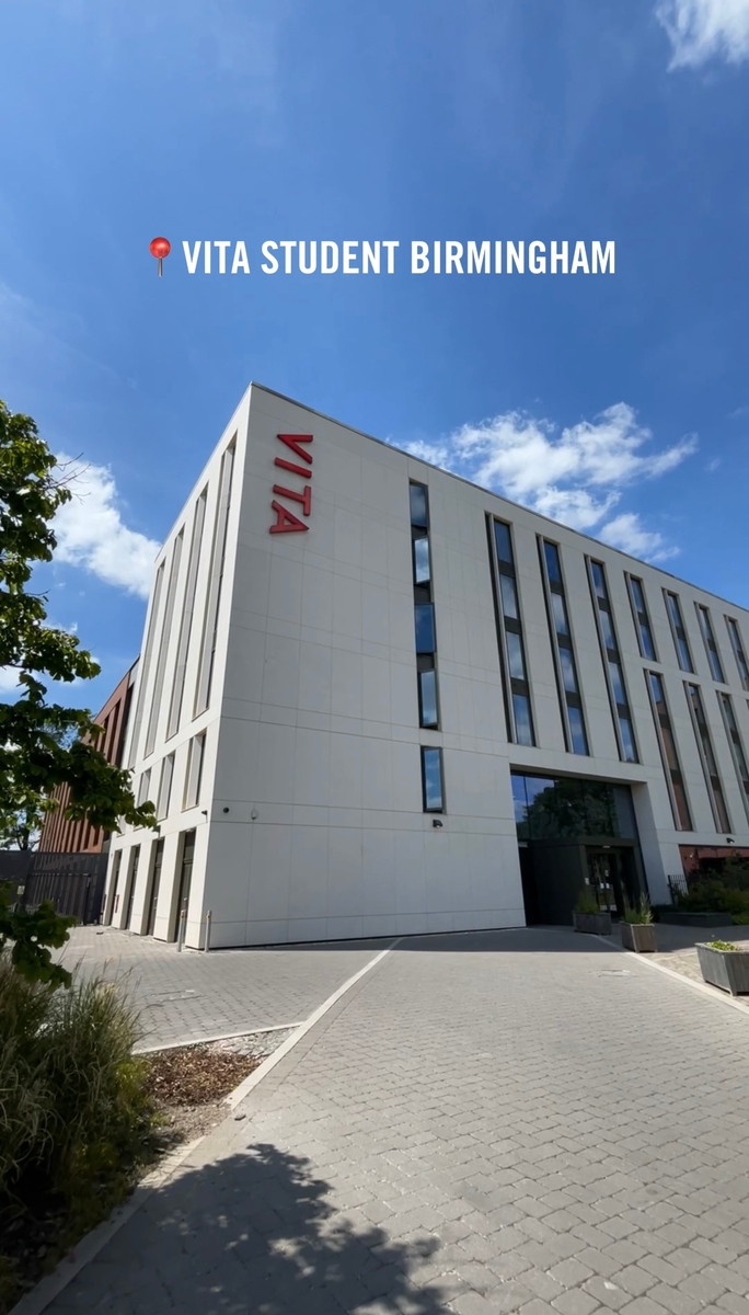 A modern, multi-story building labeled "VITA" with a pathway leading to it. The facade is mostly white with large windows. A tree and landscaped area are visible to the left. Sky above is bright and partly cloudy. Text reads "VITA Student Birmingham.