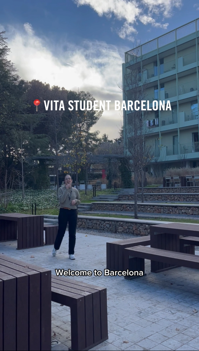 A person stands outdoors near benches, surrounded by a modern building and trees. The text reads "Vita Student Barcelona" with a red location pin above, welcoming them to student accommodation in Barcelona. The sky is partly cloudy.