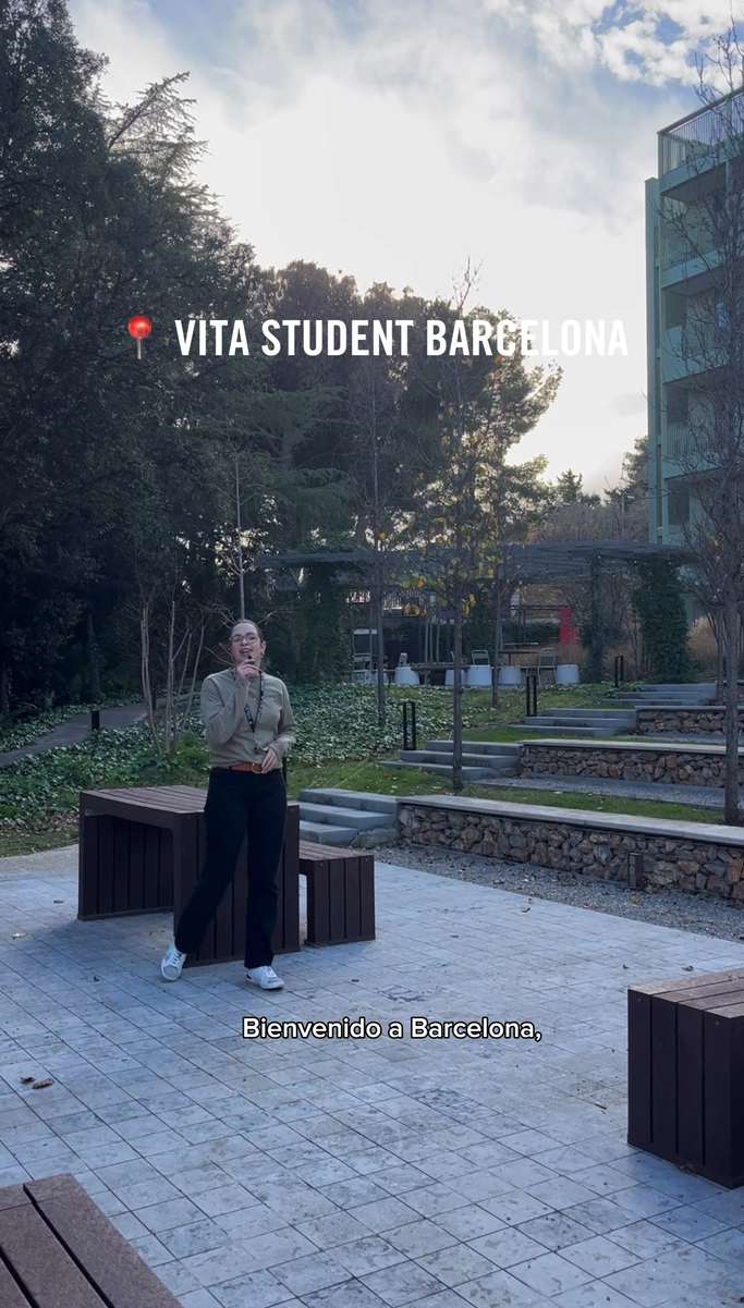 A person stands on a paved area outdoors, surrounded by trees and greenery. Text reads "VITA Student Barcelona" and "Bienvenido a Barcelona." The sky is partly cloudy, reflecting the vibrant life at this premier student accommodation in Barcelona.