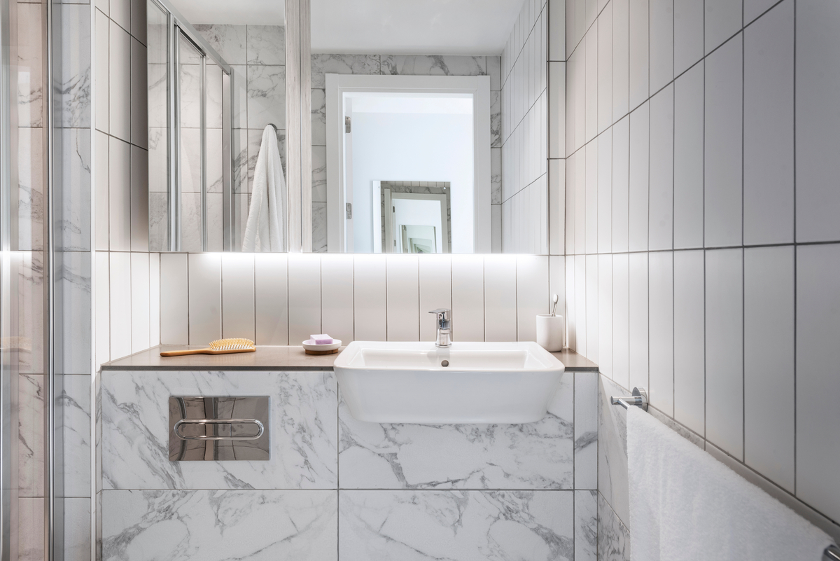 Modern bathroom with white marble tiles, a wall-mounted vanity with a sink, and a rectangular mirror. A wooden brush and soap are on the countertop. A towel hangs to the right, and a door with a large mirror is visible in the background.