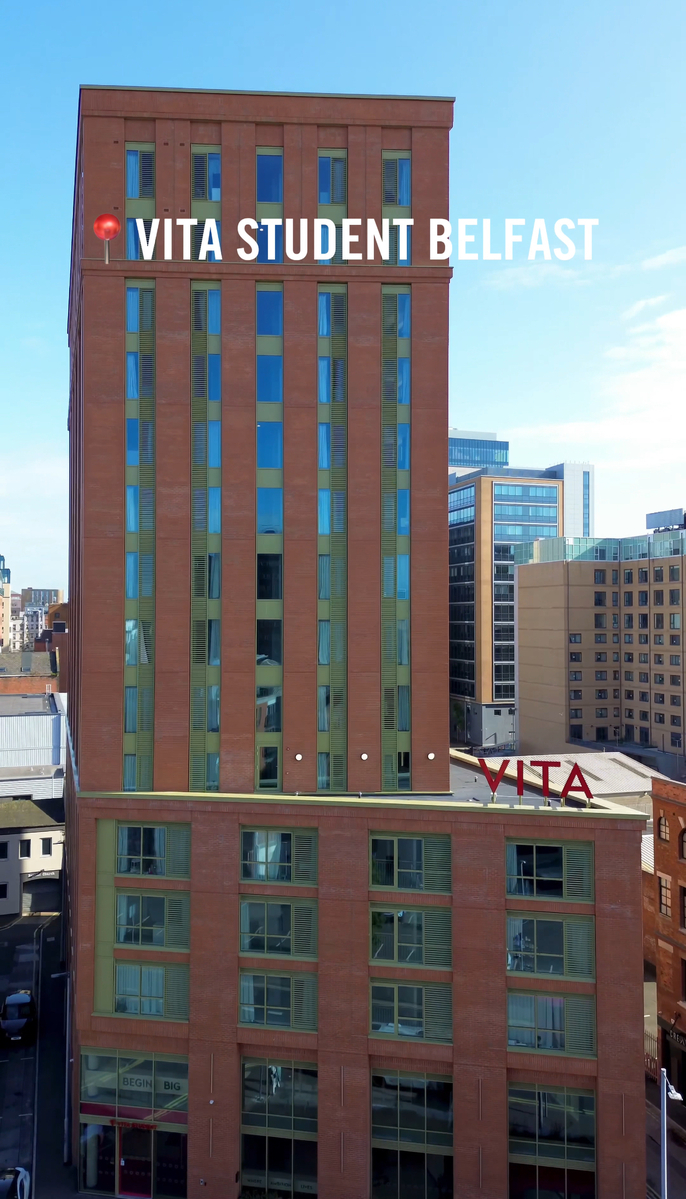 Tall red brick building labeled "Vita Student Belfast" in white text, with multiple windows across its facade. Surrounding the building are other structures, set against a clear blue sky.