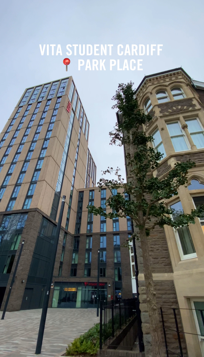 Tall modern building with "Vita Student Cardiff" sign, located on Park Place. The structure is flanked by a tree and an older architectural building with arched windows. The sky is overcast, creating a neutral backdrop.