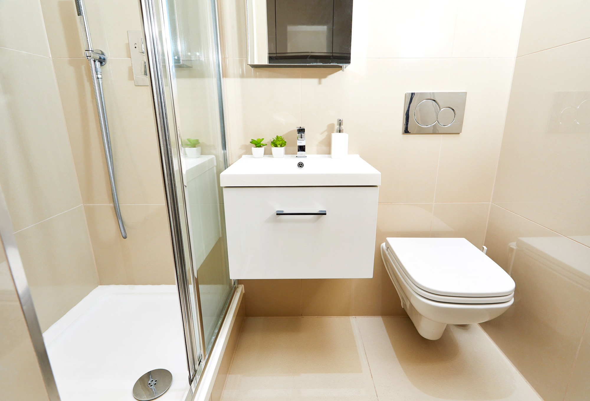 A modern bathroom in a luxury student accommodation features a glass shower enclosure on the left, a white floating vanity with a faucet and small green plants in the center, and a wall-mounted toilet on the right. The walls and floor are tiled in light beige, creating an elegant space.