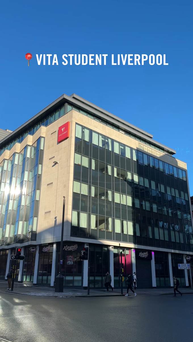 The image shows a modern, multi-story building with large glass windows under a clear blue sky. A sign reads "Vita Student Liverpool." Pedestrians are on the sidewalk below, and there's a storefront marked "Kaspa's" visible in the lower part.