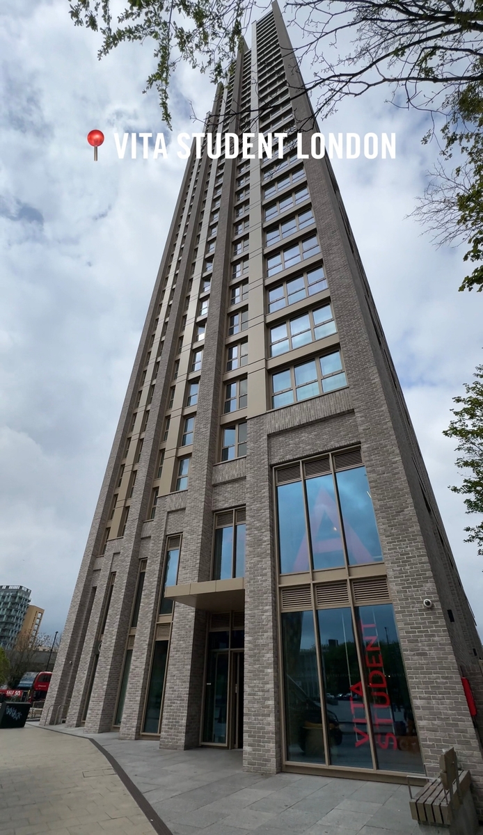 Tall modern building with a grey brick facade, numerous floors, and large windows. The entrance is visible at the bottom. Overcast sky with some trees partially framing the image. Text says "Vita Student London" near the top.