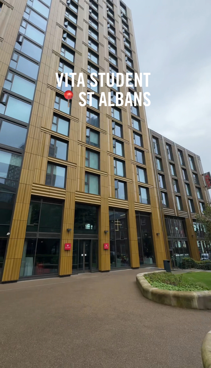 Tall modern building with wooden facade panels and large windows, labeled "Vita Student St Albans." The entrance is at the base with clear glass doors. Overcast sky and a paved area with greenery in front.