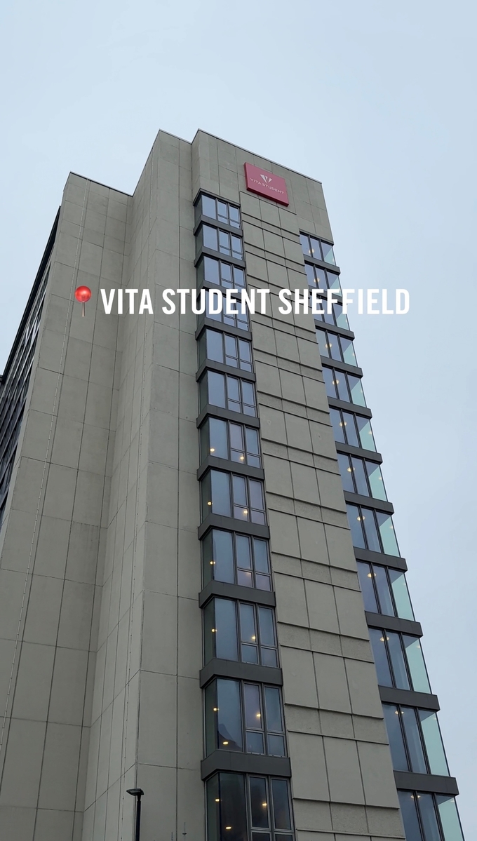 Tall building with a light gray facade, featuring large vertical windows. A bright red sign at the top reads "Vita Student," and text overlays the image: "📍VITA STUDENT SHEFFIELD" against an overcast sky background.