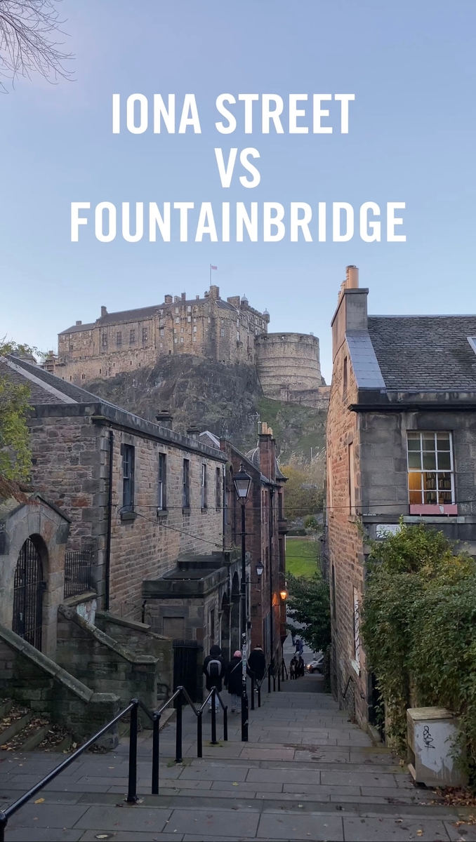 A narrow cobblestone street descends between stone buildings, leading towards a large historic castle on a hill in the background. Text at the top reads "IONA STREET VS FOUNTAINBRIDGE." Trees with autumn leaves are visible.