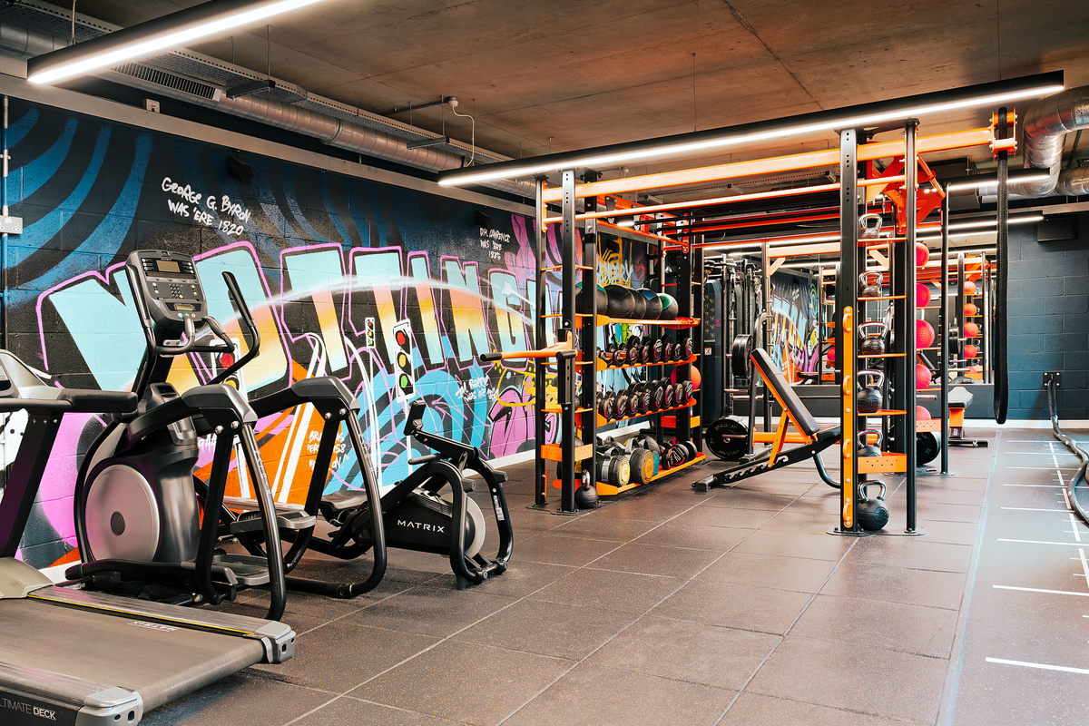 A modern gym nestled within student accommodations in Nottingham, complete with treadmills, strength training equipment, and a colorful graffiti mural. Bright lighting highlights free weights and exercise machines on a tile floor with exposed ductwork on the ceiling.