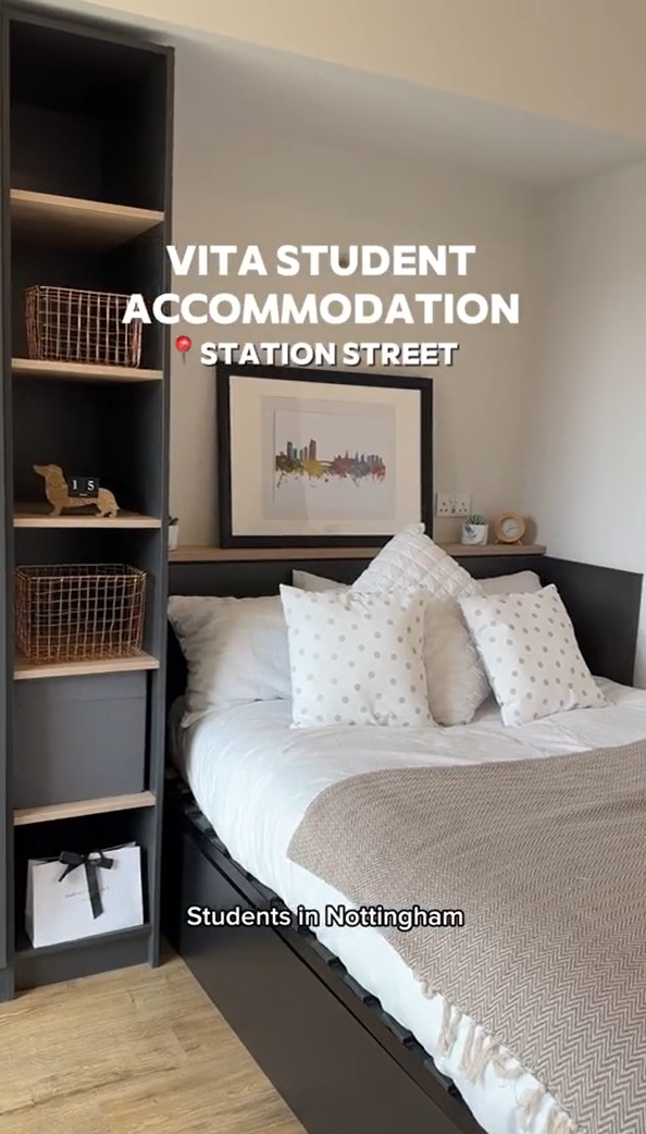 In student accommodation in Nottingham, a cozy bedroom features a neatly made bed with white and beige bedding, surrounded by throw pillows. A framed picture hangs above, while a shelving unit with baskets and a small decorative item stands beside the bed.