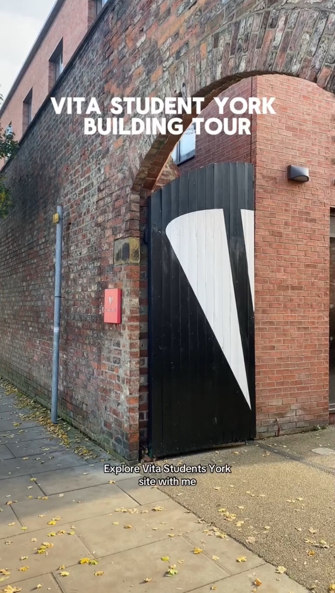 A brick building with an archway labeled "Vita Student York Building Tour" has a black door with a white design. A small red sign with "V" is on the wall. Leaves are scattered on the sidewalk. Text on the image says "Explore Vita Student York site with me.