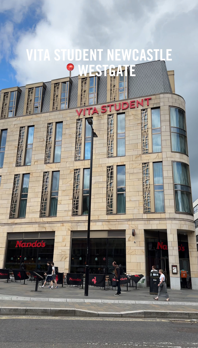 Exterior view of Vita Student Newcastle Westgate building, featuring large windows and a Nando's restaurant on the ground floor. People are walking on the sidewalk in front. The sky is partly cloudy.