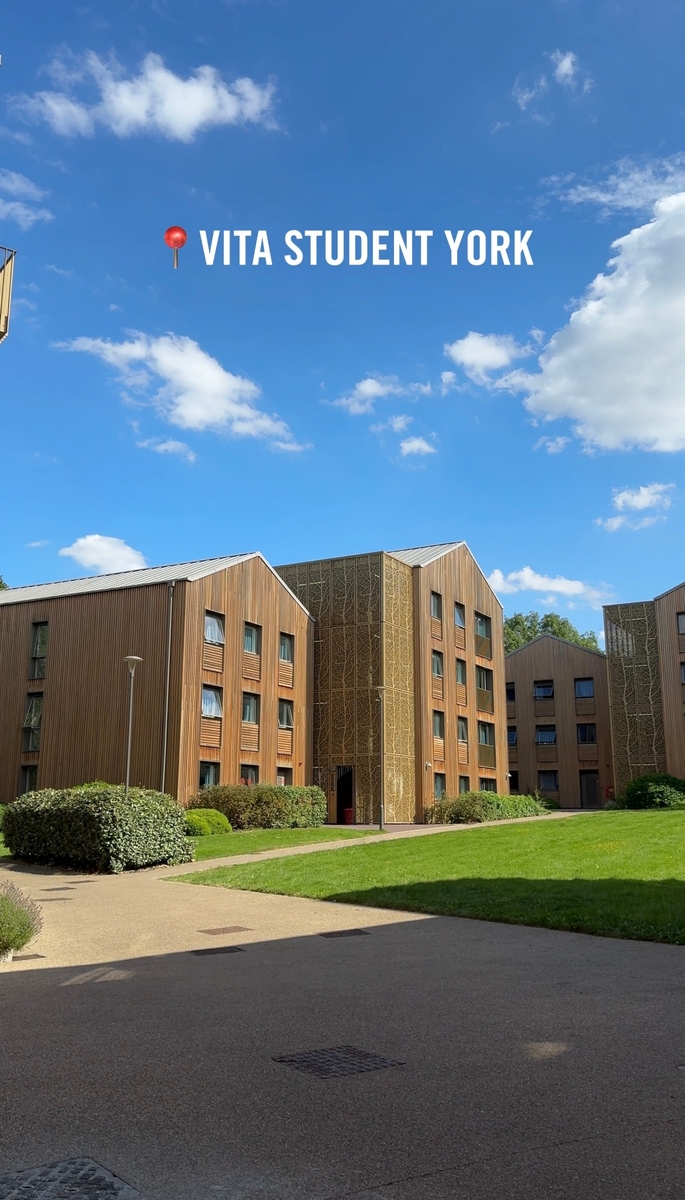 Modern wooden student apartments with large windows, set in a green, grassy area under a bright blue sky with a few clouds. Text at the top reads, "Vita Student York.