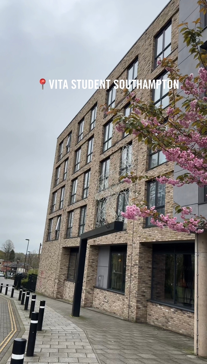 A modern brick building with large windows stands under a cloudy sky. In the foreground, a cherry blossom tree adds charm to Portswood House. A sign reads "VITA STUDENT SOUTHAMPTON." Bollards neatly line the sidewalk.