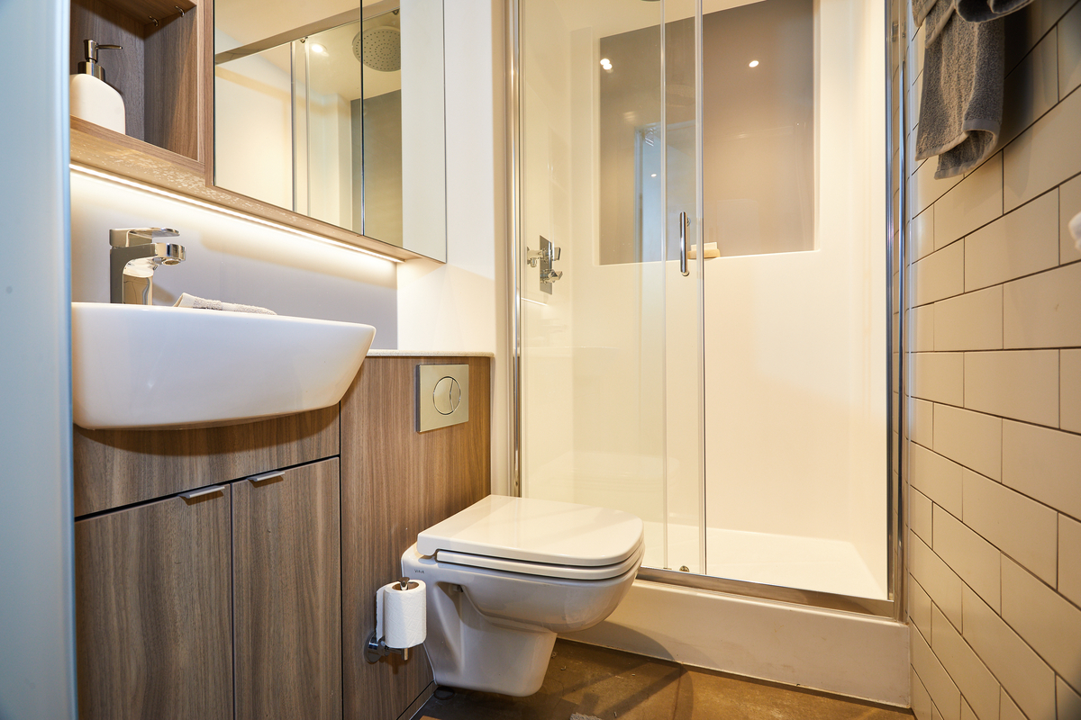 Modern bathroom in Portswood House featuring a white sink on a wooden cabinet, wall-mounted toilet, and glass-enclosed shower. The white-tiled walls and warm lighting enhance the clean, contemporary look.