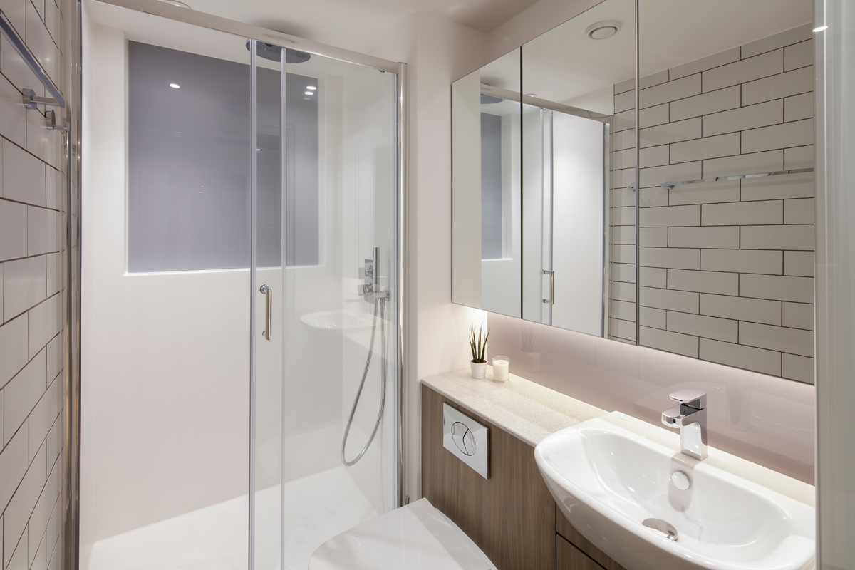 A modern bathroom in the heart of Manchester features a glass shower enclosure, white subway tiles, and a wooden vanity. A white basin with a chrome faucet accents the room, accompanied by a toilet. A mirror reflects the space while a small plant adds life to the countertop.