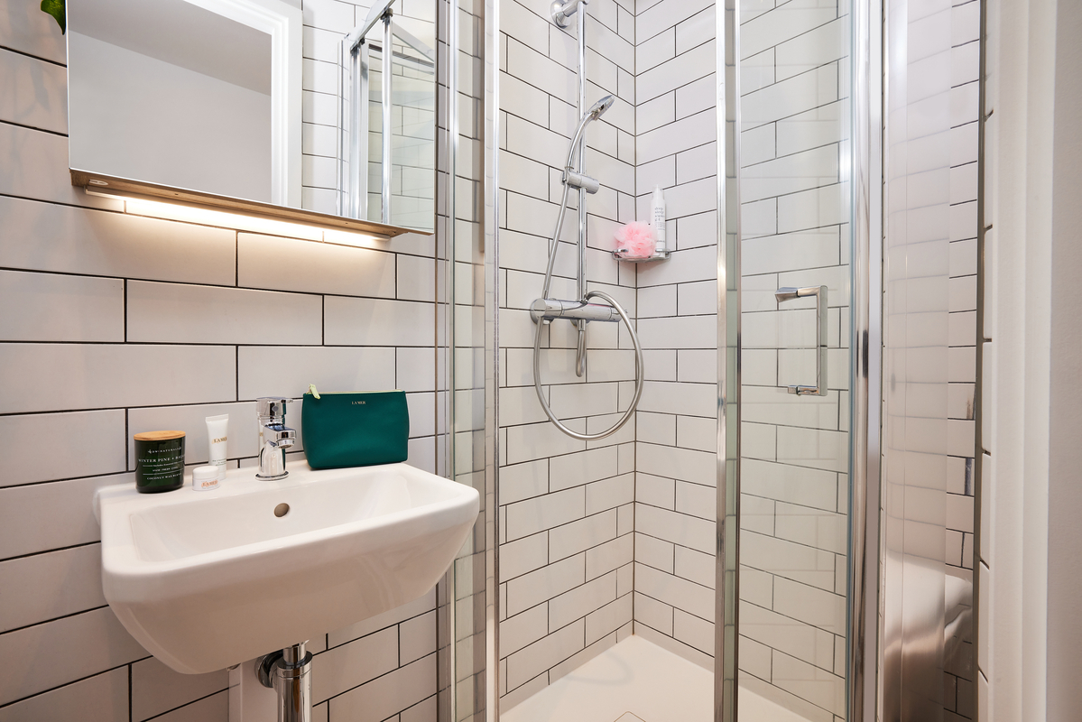 Modern bathroom with white subway tile walls, a small corner sink with toiletries, a glass-enclosed shower, and a showerhead. A green pouch and a pink shower pouf are on display.