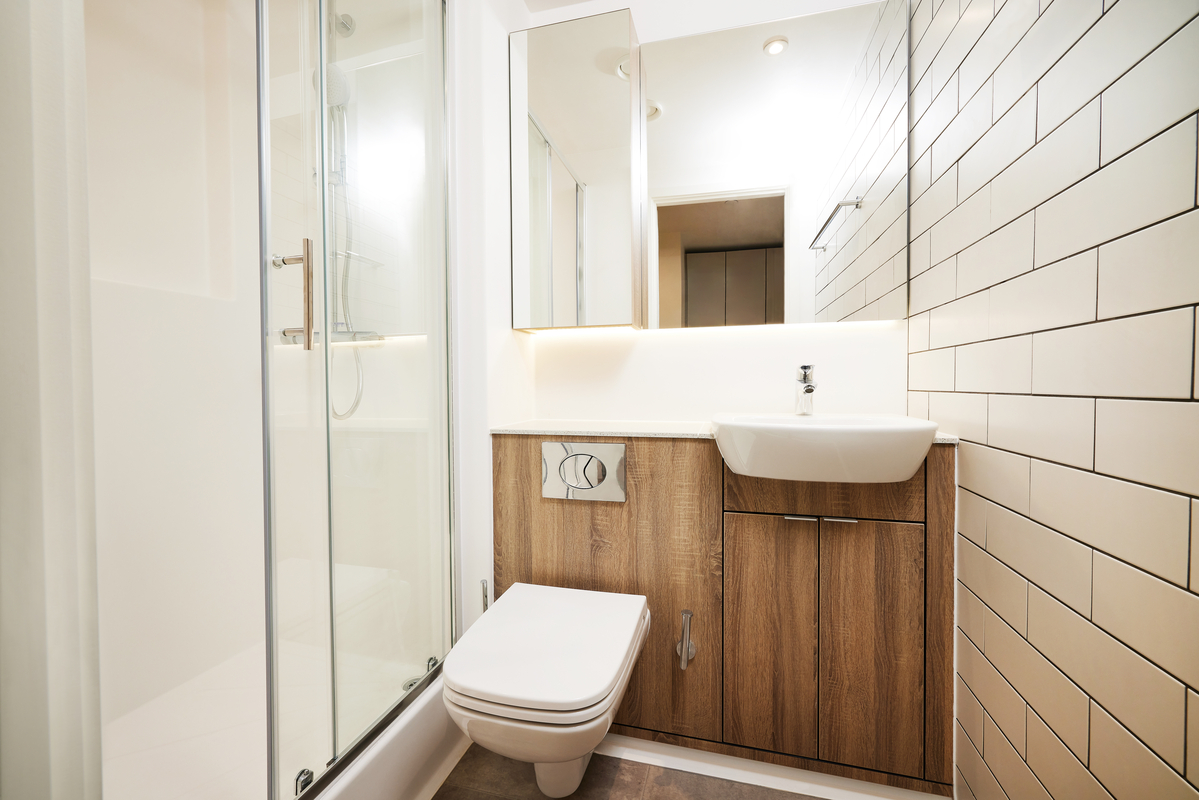 This modern bathroom in the heart of Belfast boasts beige tiled walls, a sleek glass shower enclosure, and a wall-mounted toilet. The wooden vanity with a white sink complements its minimalist style, creating an oasis of tranquility in the bustling city atmosphere.