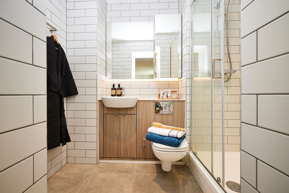 A modern bathroom with white subway tiles, a glass-enclosed shower, a wood-paneled sink cabinet, and a wall-mounted toilet. Towels and toiletries are neatly arranged, and a black robe hangs on a hook. The floor is tiled in a neutral tone.