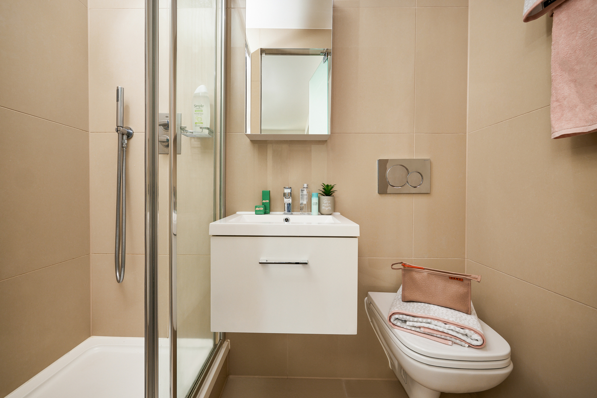 A modern city bathroom with beige tiles features a glass shower on the left, a white floating sink with toiletries, and a square mirror above. A wall-mounted toilet on the right has a pink bag and folded towel neatly placed on top. Perfect for urban living in Sheffield.