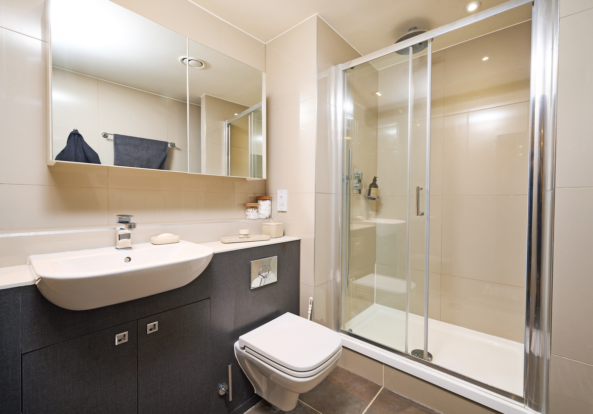 A modern bathroom with beige walls features a large mirror above a white sink with a chrome faucet. Beside it is a toilet with a wall-mounted flush. A glass-enclosed shower is on the right, with toiletries on the shelf.