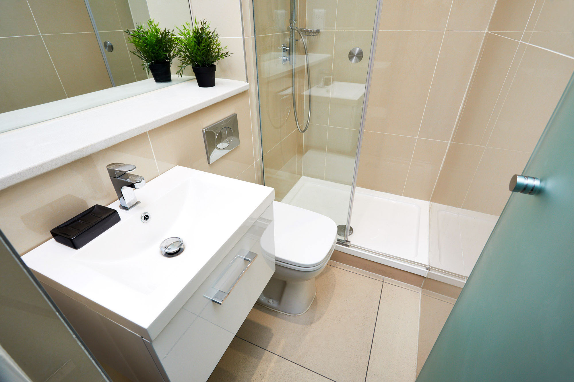 In the heart of Bristol, discover a modern bathroom featuring a white vanity sink with a silver faucet and an elegant mirror above. Next to the toilet sits a glass-enclosed shower with a metal showerhead. Two potted plants on the countertop add a touch of greenery to this urban oasis.