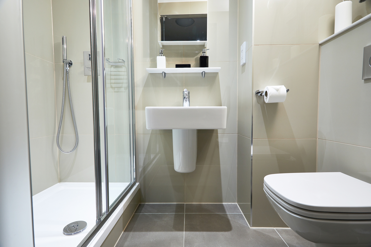 Modern bathroom with beige tiles, reminiscent of a stylish Glasgow apartment, featuring a glass-enclosed shower on the left, a white sink with a mirror and shelf above in the center, and a wall-mounted toilet on the right. A toilet paper holder is mounted on the wall.