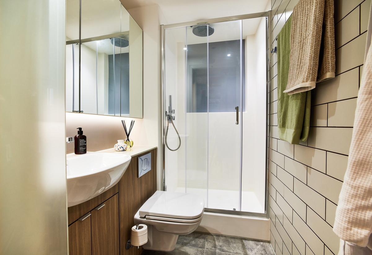 In the heart of Manchester, this modern bathroom boasts a glass-enclosed shower with a rain showerhead. It features a wall-mounted sink atop a wooden cabinet, with a mirror above. Neutral-toned tiles cover the floor and walls, complemented by towels hanging on a rail.