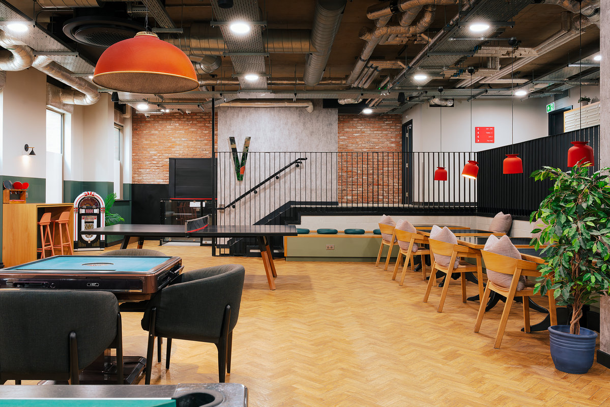 Spacious modern game room with ping pong table, card table, and wooden dining chairs. Industrial design features exposed brick, ceiling pipes, and bright red hanging lamps. A large plant decorates the corner, enhancing the cozy atmosphere.