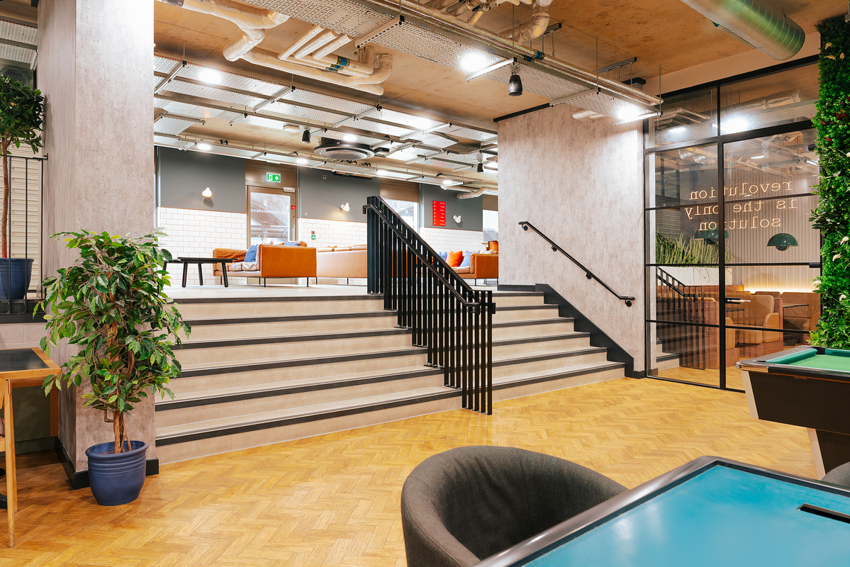 Modern office lounge with a staircase leading to a seating area with orange couches. A potted plant is on the left, and pool tables are in the foreground. Walls have industrial decor with exposed pipes, and glass partitions.