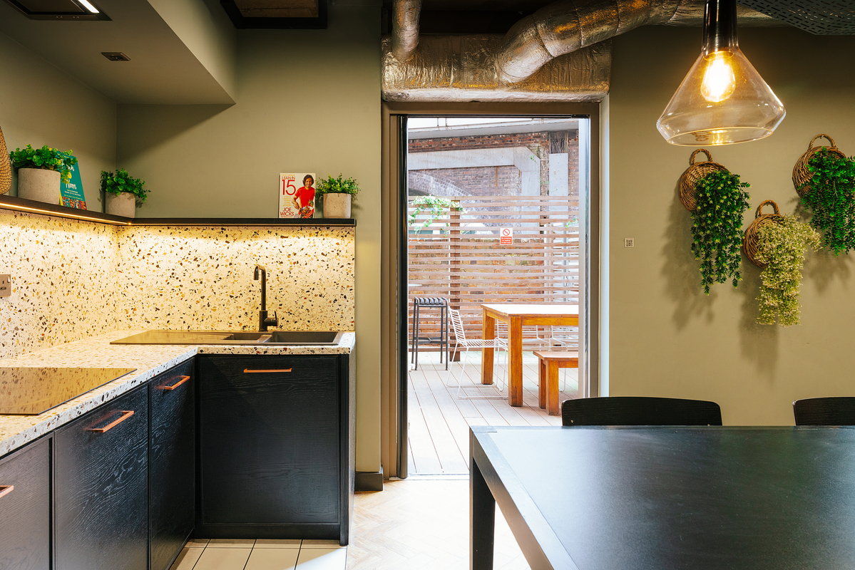 Modern kitchen with black cabinets, speckled countertop, and overhead lighting. Potted plants decorate the walls. Large glass door opens to a wooden patio with a table and chairs, bordered by a wooden privacy screen.