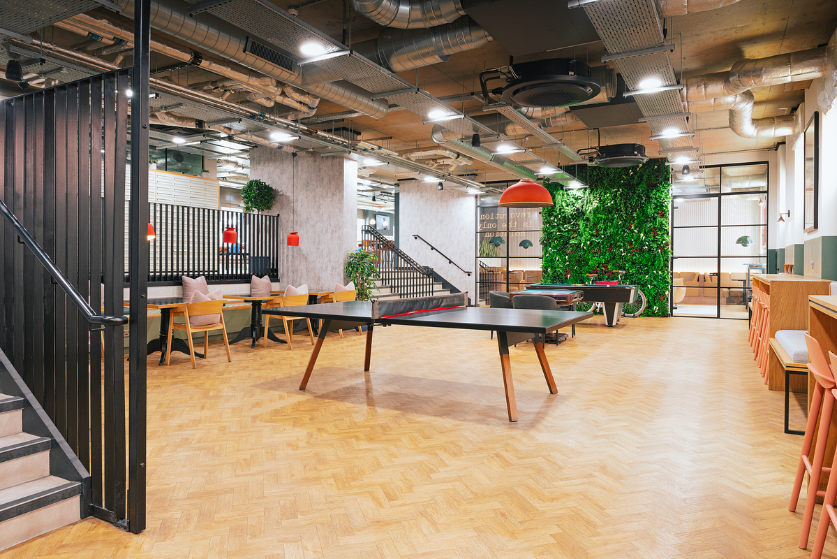 A modern office space featuring a ping pong table, a seating area with cushions, a wall with greenery, and high chairs along a counter. The ceiling exposes ductwork, and the room is lit by industrial-style fixtures.