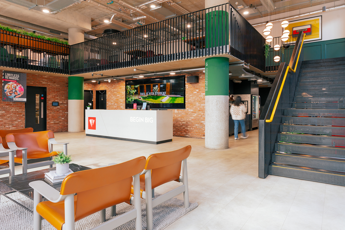 Modern office lobby with brick walls and green pillars. Features an orange seating area, a reception desk displaying "Begin Big," and a staircase leading to an upper level. Plants and art pieces add decor. Two people are near the reception area.