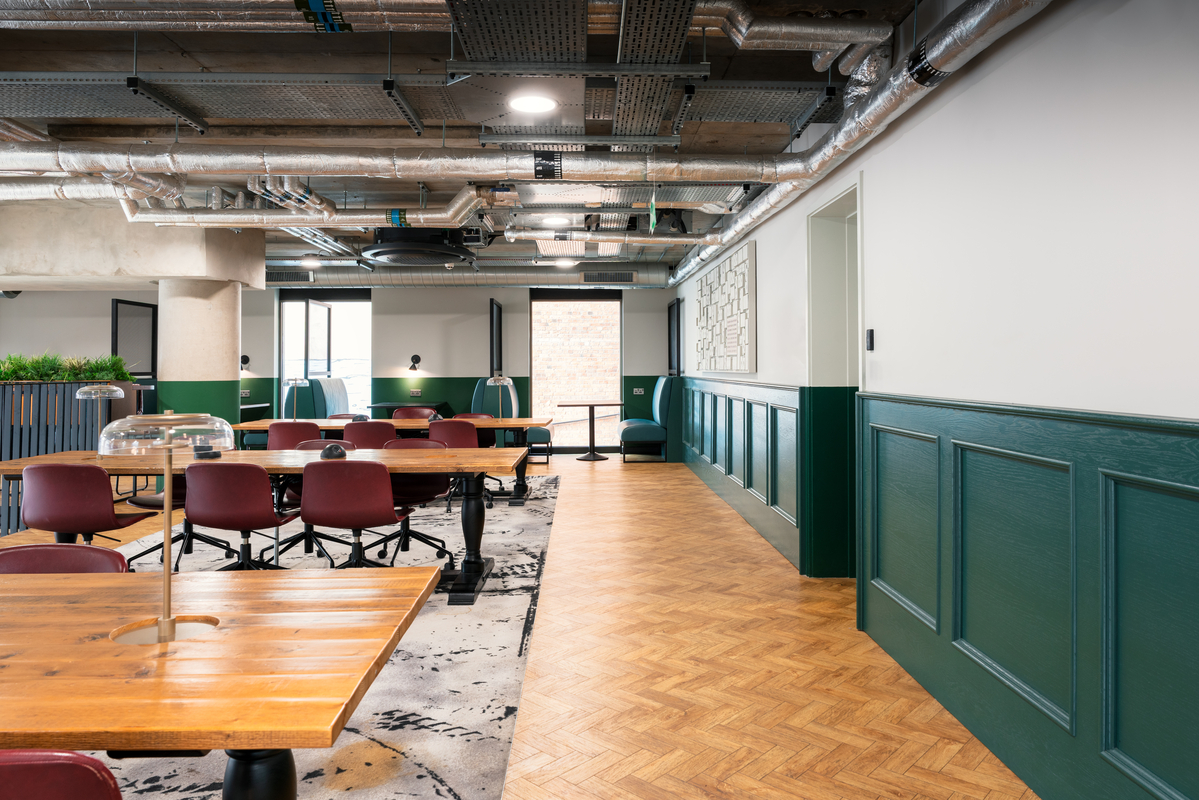 A modern office space with wooden tables, red chairs, and a patterned rug. Green panels line the white walls, and a large window lets in natural light. Exposed ceiling pipes and sleek lighting fixtures add an industrial touch.