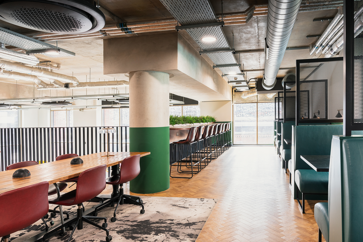 Modern office space with wooden flooring, a long wooden table with red chairs, and high stools by a bar-style counter. Exposed ductwork and large pillars accent the industrial design. Natural light enters through large windows.