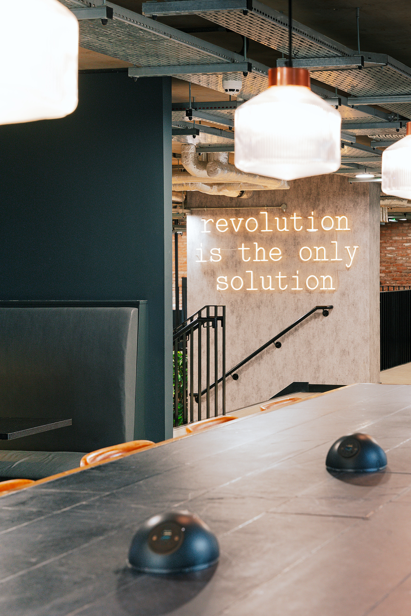 Modern interior with a black table and overhead lights. A neon sign on the wall reads "revolution is the only solution." Stairs and a brick wall are in the background.