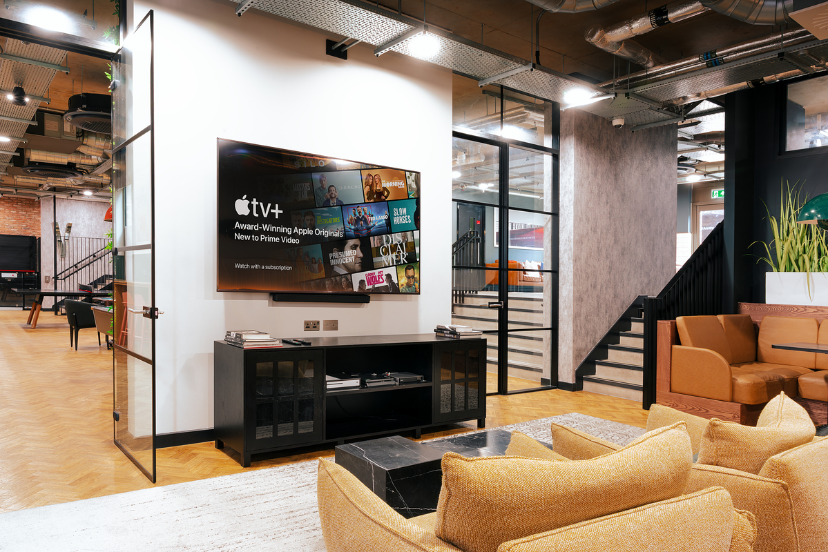 A modern office lounge featuring a TV displaying Apple TV+ content on a white wall. There's a black cabinet under the TV, a gray sofa set with mustard cushions, and an open space with stairs, plants, and colorful chairs in the background.