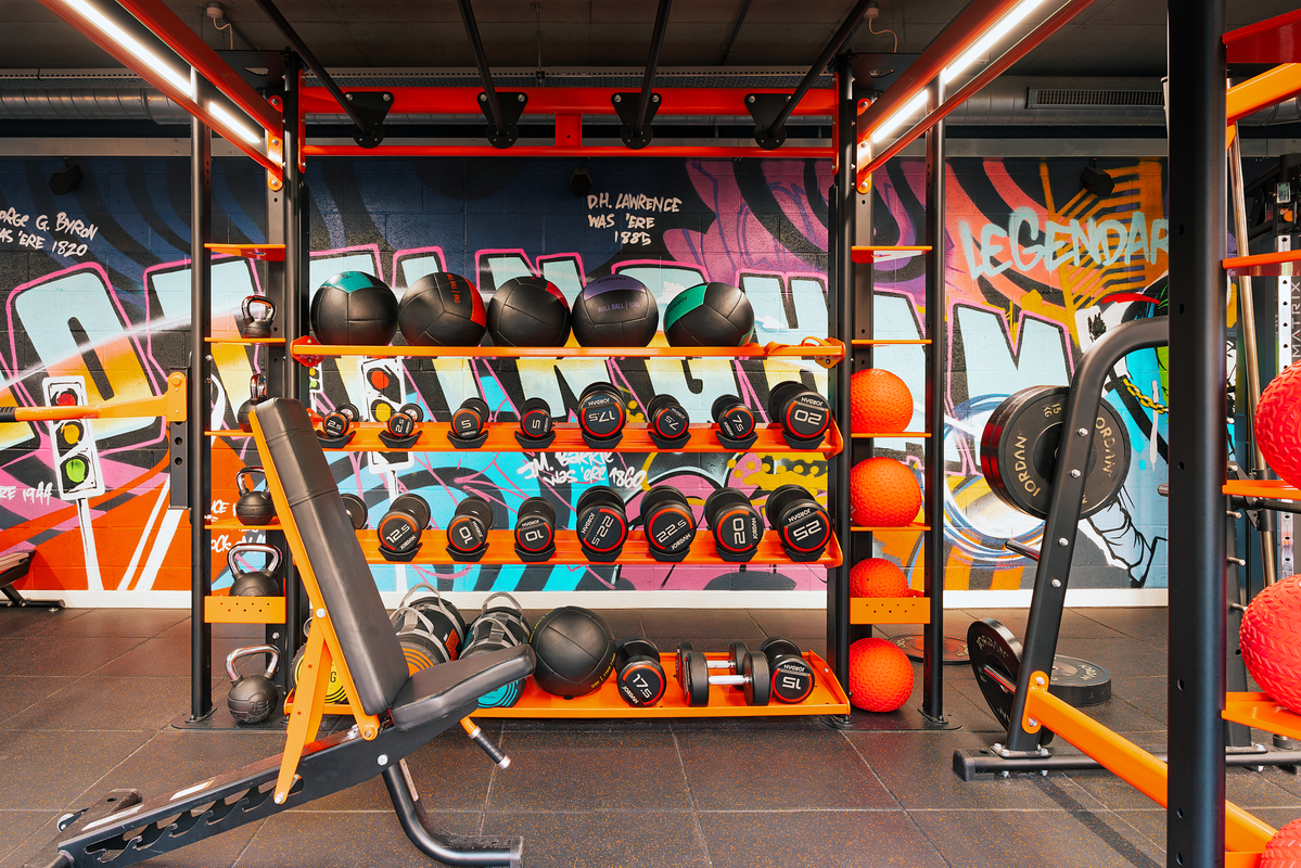 Gym with colorful graffiti, weightlifting equipment, and a bench. Shelves hold kettlebells, dumbbells, medicine balls, and other gear. Brightly lit with an energetic atmosphere.