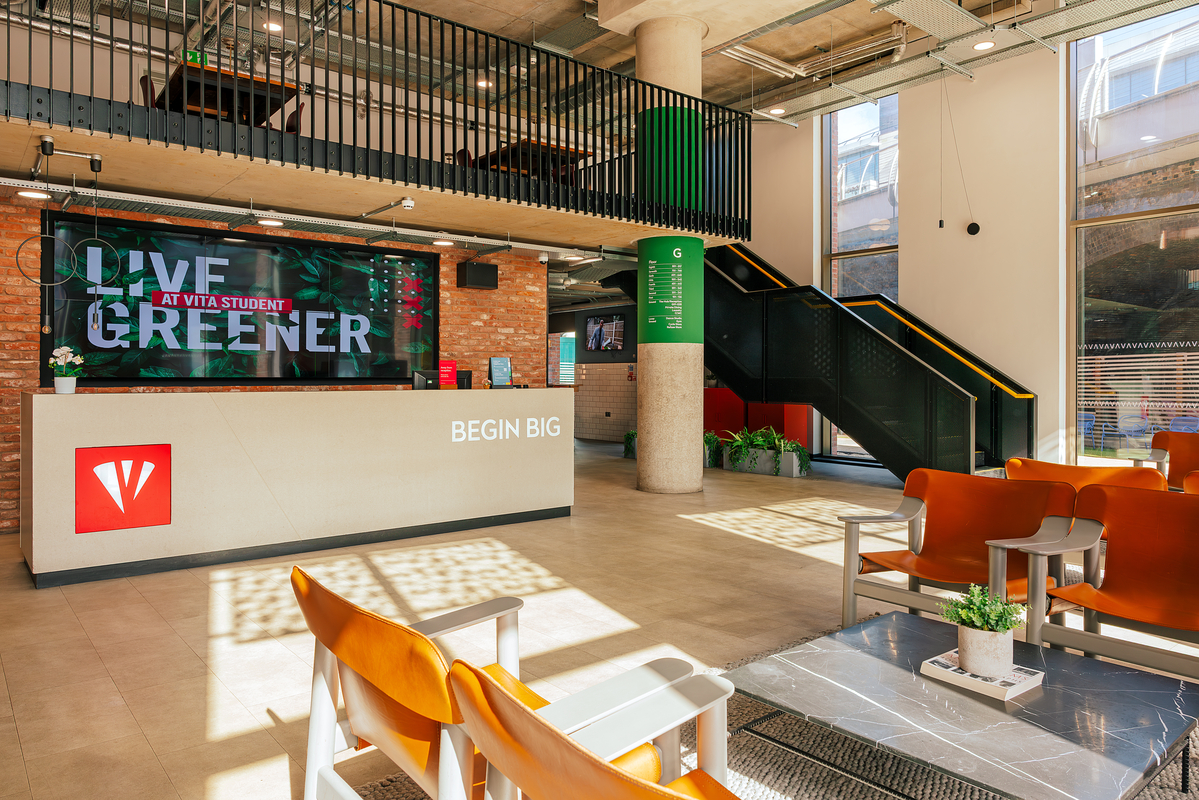 Bright, modern lobby with a reception desk displaying "Live Greener." Wooden chairs are arranged around tables with plants. A staircase and balcony with industrial design elements are visible. Large windows allow natural light inside.