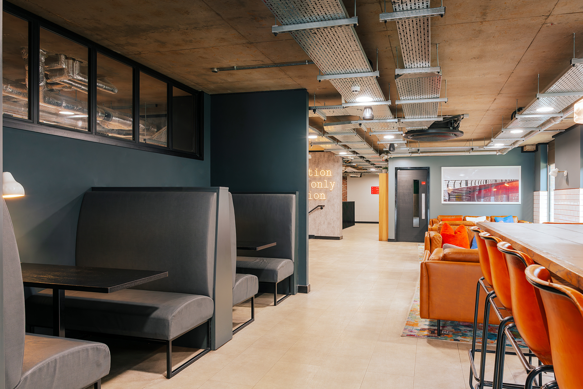 Modern office space with black booths and a long wooden table with orange chairs. Exposed pipes and ducts are visible on the ceiling. The walls are dark with a mix of industrial and cozy design elements. A framed photo is mounted on the far wall.