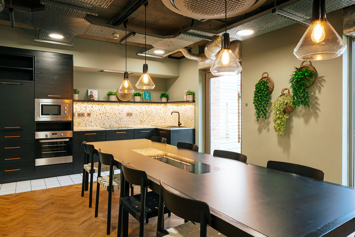 A modern kitchen features a dark central table with chairs, hanging pendant lights, and an array of sleek cabinets. The backsplash is speckled, and the walls are decorated with potted plants and woven baskets, creating a cozy, contemporary ambiance.