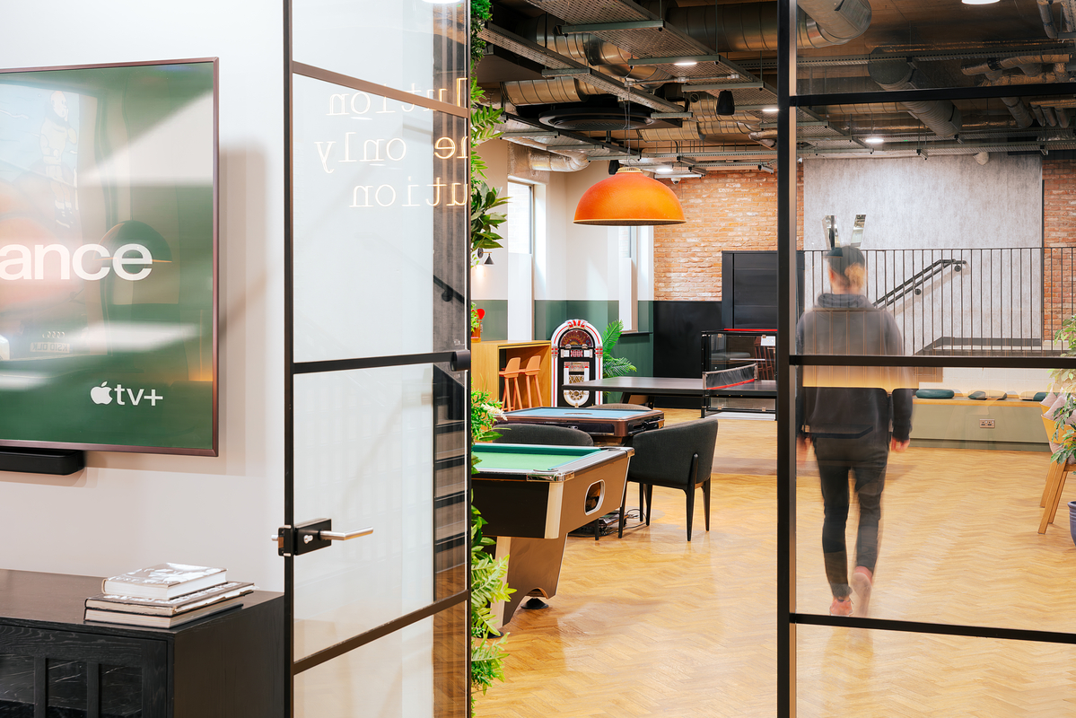 A bright room with glass doors showing a game area. Inside, there's a pool table, jukebox, and orange pendant light. A person walks away, facing a seating area with a TV on the wall, displaying Apple TV+.