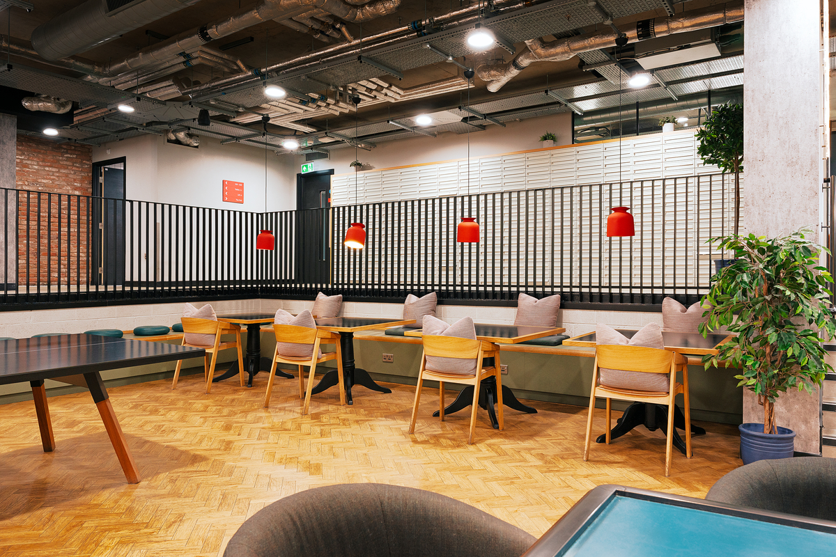 Modern workspace with wooden flooring, tables, and chairs. Red pendant lights hang above a long bench with cushions. Exposed ceiling pipes and brick walls add an industrial touch. Potted plant and natural light enhance the space.