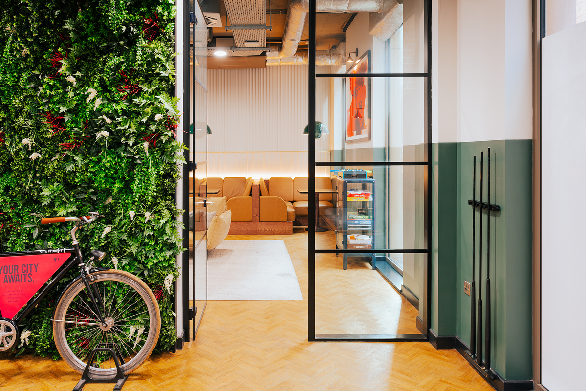 Office entrance with a black-framed glass door, wood flooring, and a vertical garden wall with a bicycle. Inside, a cozy seating area features a warm-toned sofa and chairs, under soft lighting and modern decor.